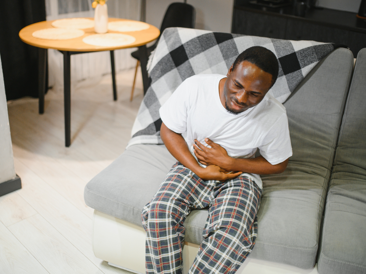 A man thinking about what to take for stomach bug, in plaid pajama pants and a white t-shirt sits on a couch, holding his stomach and wincing in pain. A checkered blanket is draped over the couch, and a dining table is visible in the background.