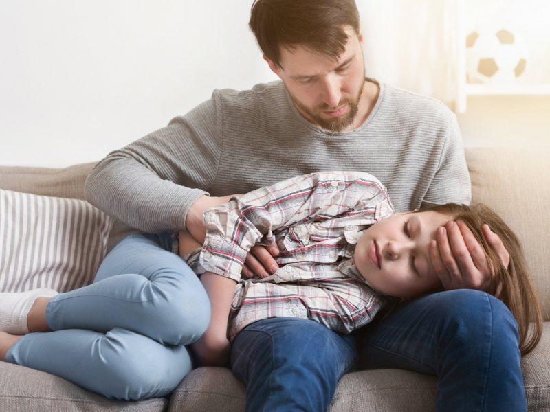 A man with a stomach bug going around​, sits on a couch holding and comforting a young girl who appears unwell, resting her head on his lap as he gently touches her forehead. A soccer ball is visible in the background.