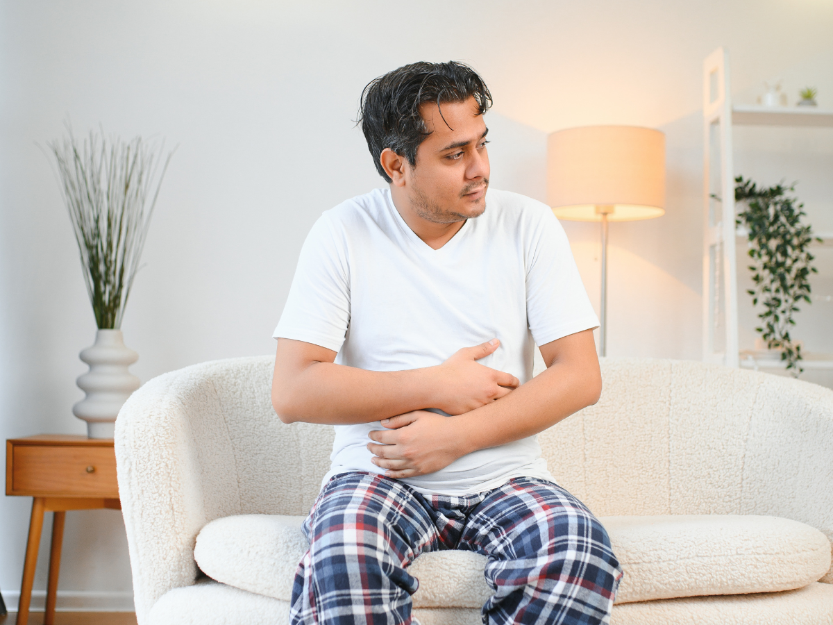 A man with rebound constipation in pajamas sits on a white couch, holding his stomach and appearing uncomfortable, possibly experiencing abdominal pain. The room is softly lit with a lamp and decorated with plants.