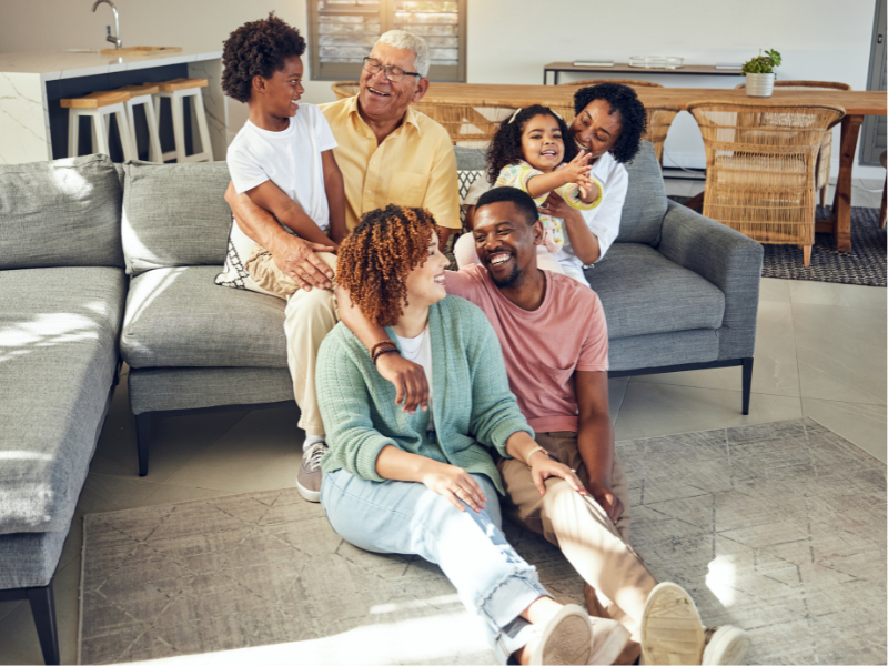 A multi-generational family of six relaxes and smiles together in a bright living room, discussing topics like the effect of antibiotics on gut flora. The joyful, warm atmosphere brings everyone closer, seated on a gray sofa or gathered on the floor.