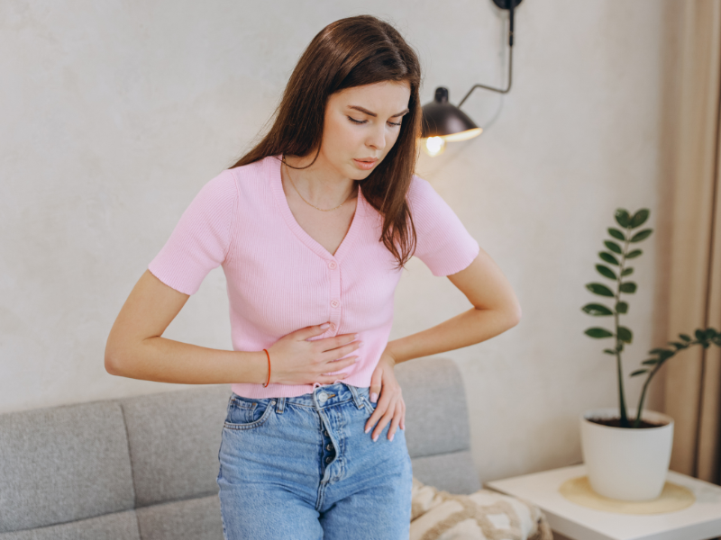 A young woman in a pink shirt and jeans stands indoors, holding her stomach with a pained expression, possibly experiencing antibiotic associated diarrhoea. A potted plant and sofa are visible in the background.