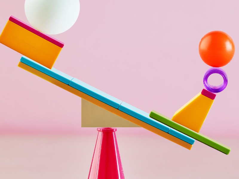 Colorful geometric shapes and balls are carefully balanced on a seesaw against a pink background, visually symbolizing balance and equilibrium—much like maintaining health and avoiding dysbiosis symptoms.